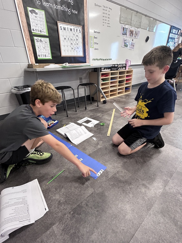 Two boys collaborate on a fraction activity, arranging paper pieces along a blue strip with rulers and worksheets nearby.