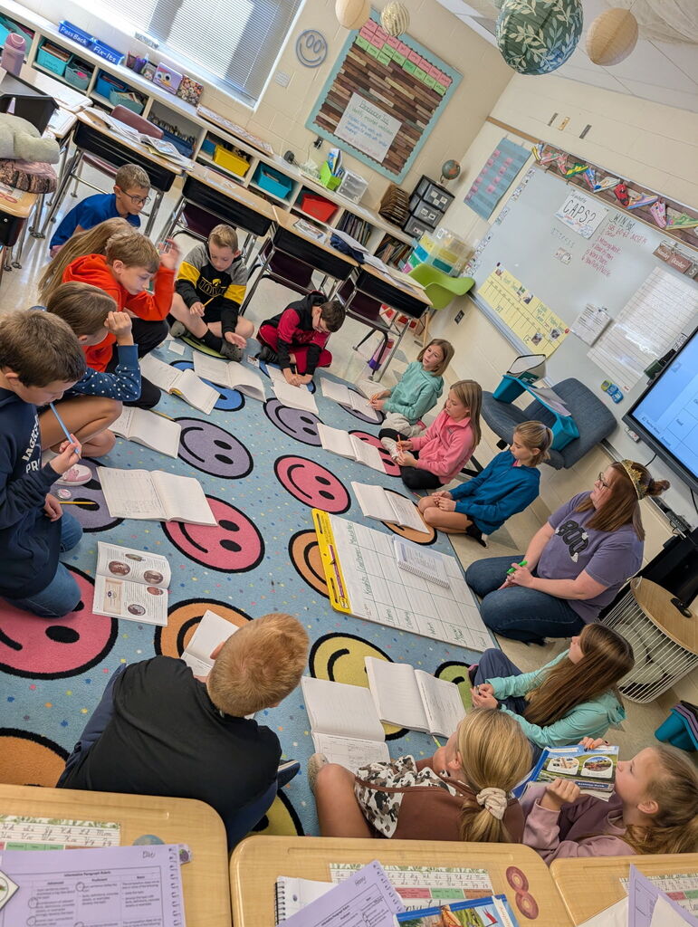 Students and a teacher sit in a circle on a colorful smiley-face rug, working in notebooks during a classroom lesson.