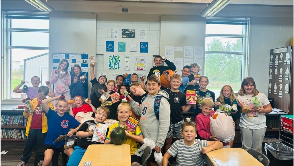A cheerful group of students poses in a classroom, proudly displaying pumpkins, crafts, baked goods, and other market purchases.