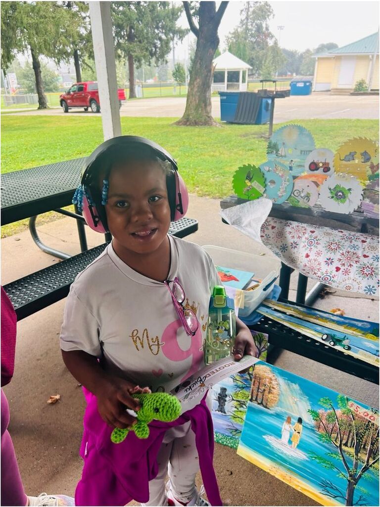A smiling student wearing pink headphones holds a crocheted green turtle while standing beside colorful artwork at an outdoor market.