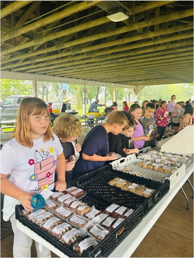Students browse tables filled with packaged baked goods and treats under a covered pavilion at a school market.