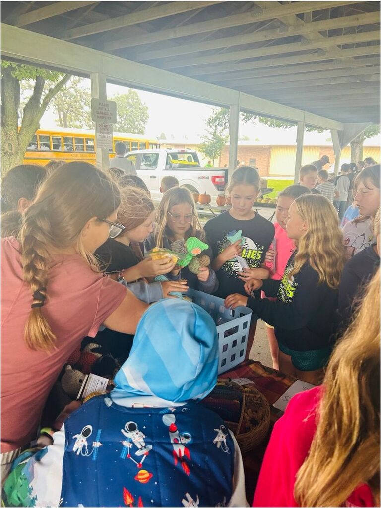 A group of students gathers around a table, examining colorful handmade crocheted animals at an outdoor school market.