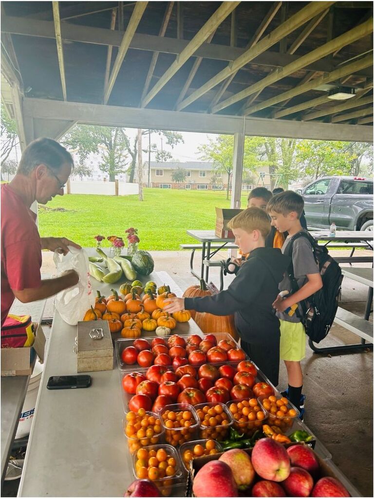 Students select miniature pumpkins and gourds from a table displaying fresh apples, tomatoes, and other produce.
