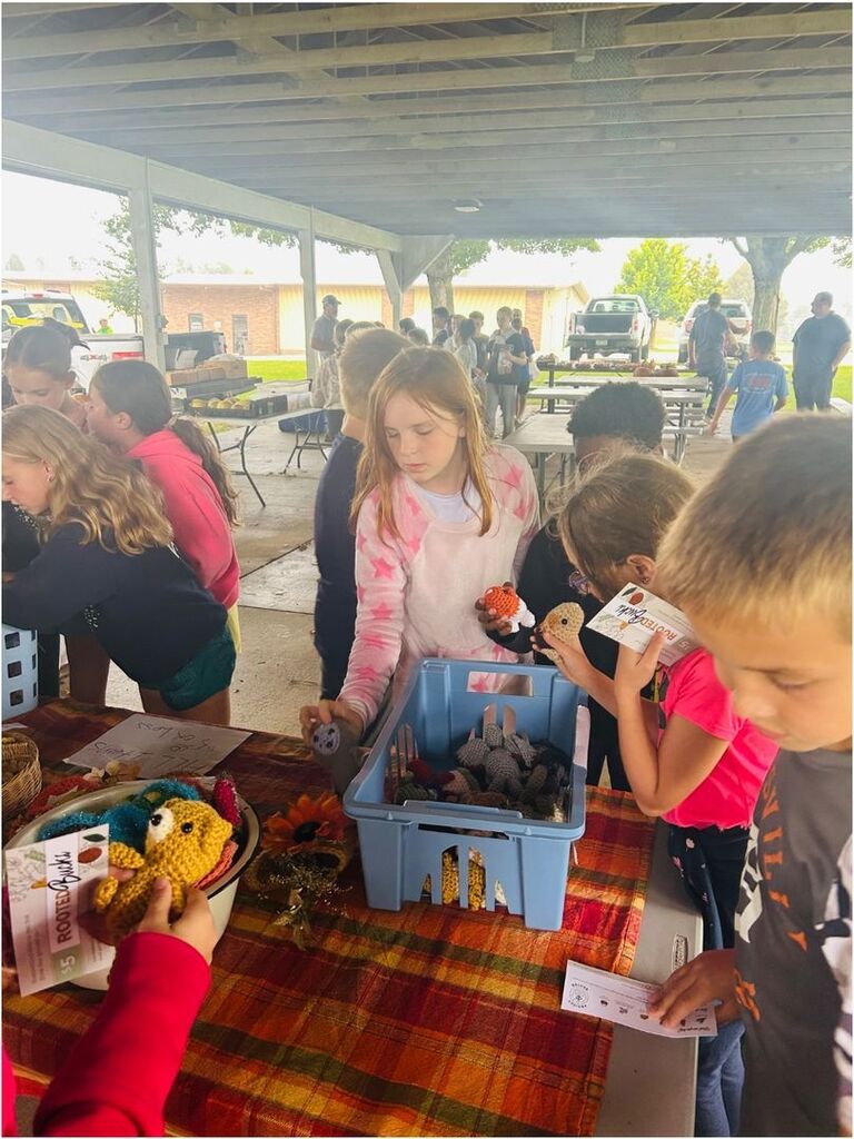Students browse handmade crocheted animals and crafts displayed in bins and baskets at a bustling outdoor school market.