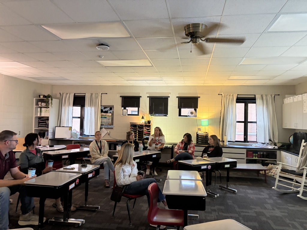 Group of adults sitting behind student desks having a conversation.