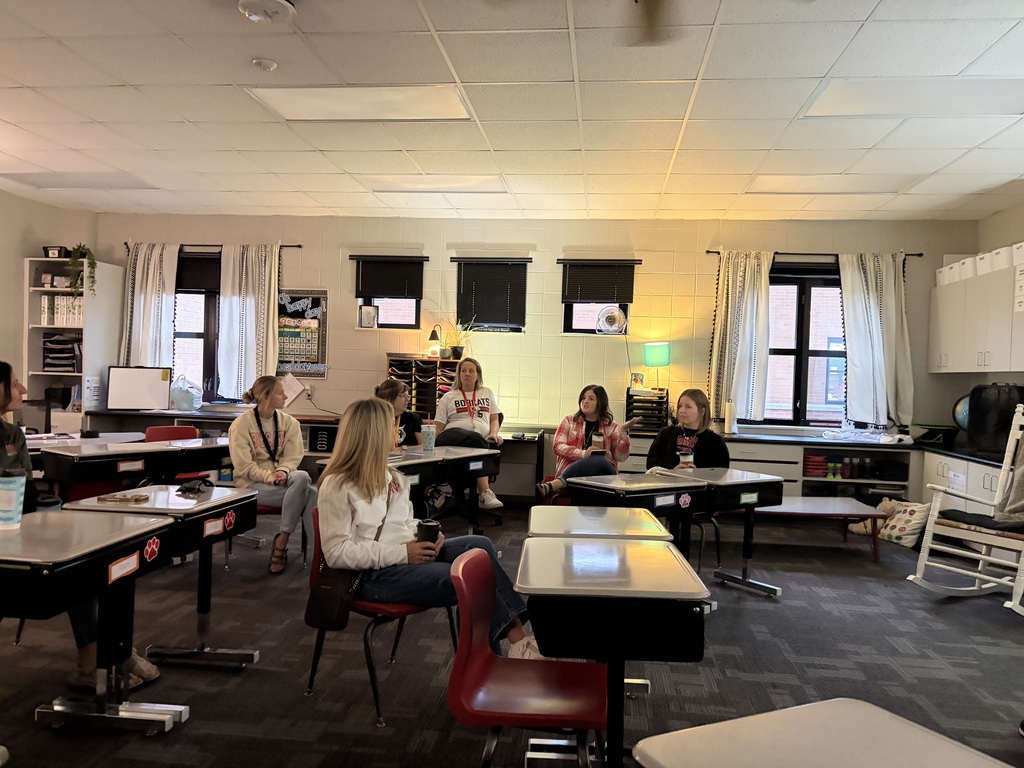 Group of adults sitting behind student desks having a conversation.