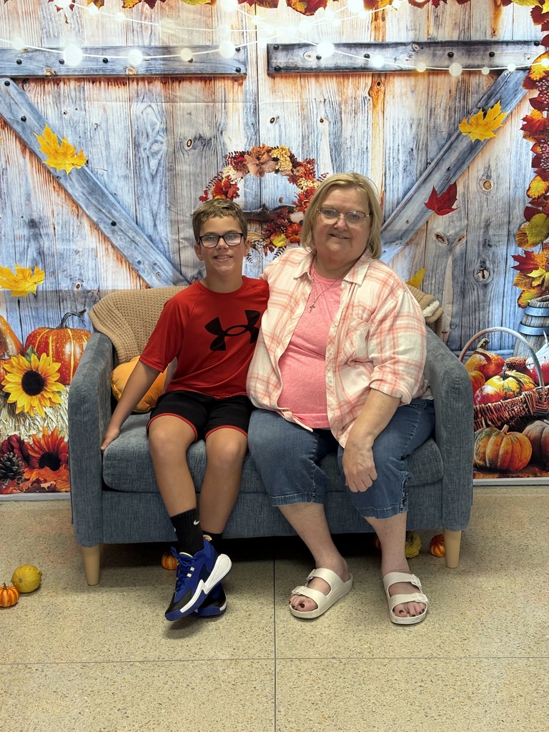 A smiling boy and woman sit together on a blue loveseat in front of a festive autumn backdrop with pumpkins and leaves.