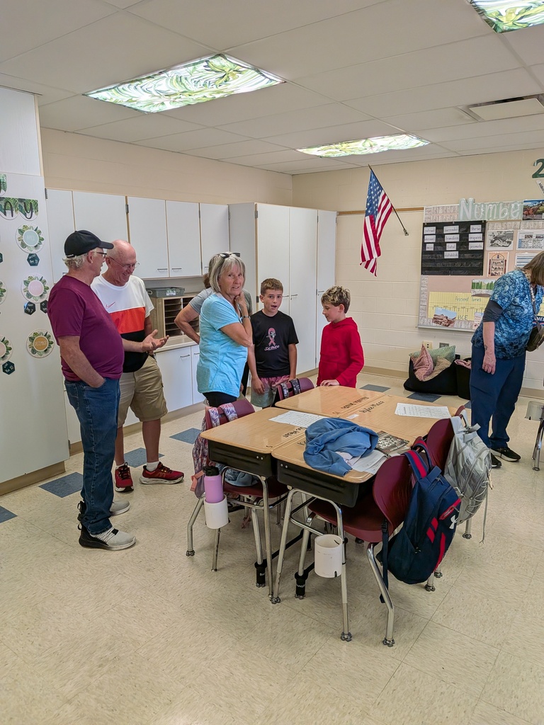 Two boys and several adults gather around desks and talk in an elementary classroom decorated with learning materials.