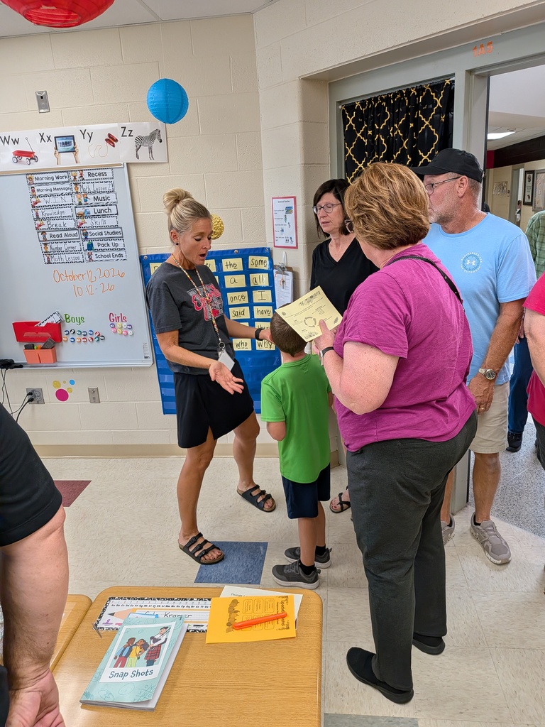 A teacher speaks with a young boy and visiting adults in an elementary classroom during a school visit.