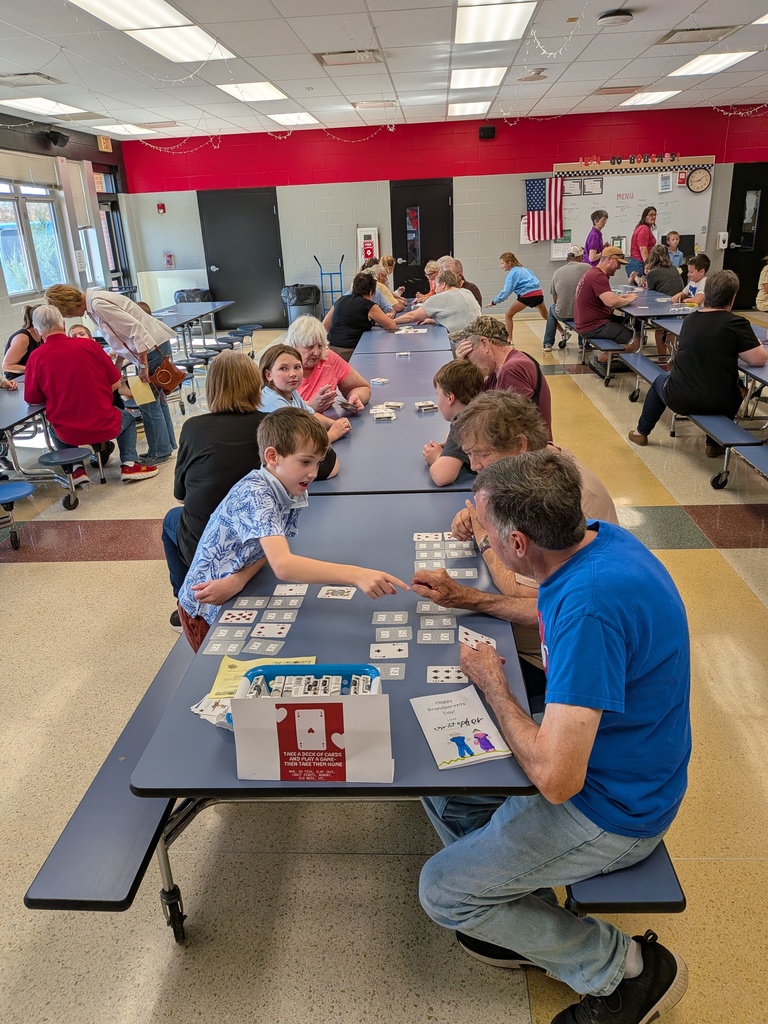 Children and older adults play card games together at cafeteria tables during a school community event.