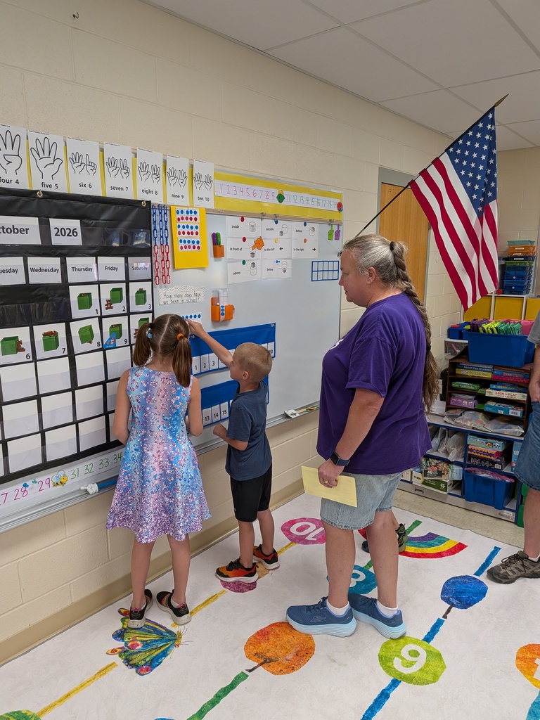 Two young students interact with a classroom calendar while an adult watches beside them.