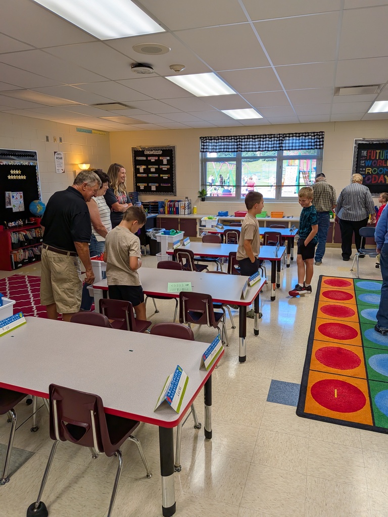 Children and adults explore an elementary classroom filled with student desks, books, and colorful decorations.