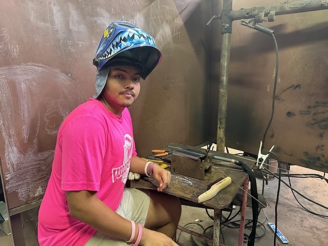 A male student wearing protective welding gear smiles  for the camera.