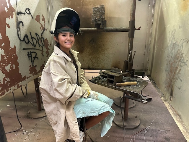 A female student wearing protective welding gear smiles  for the camera.