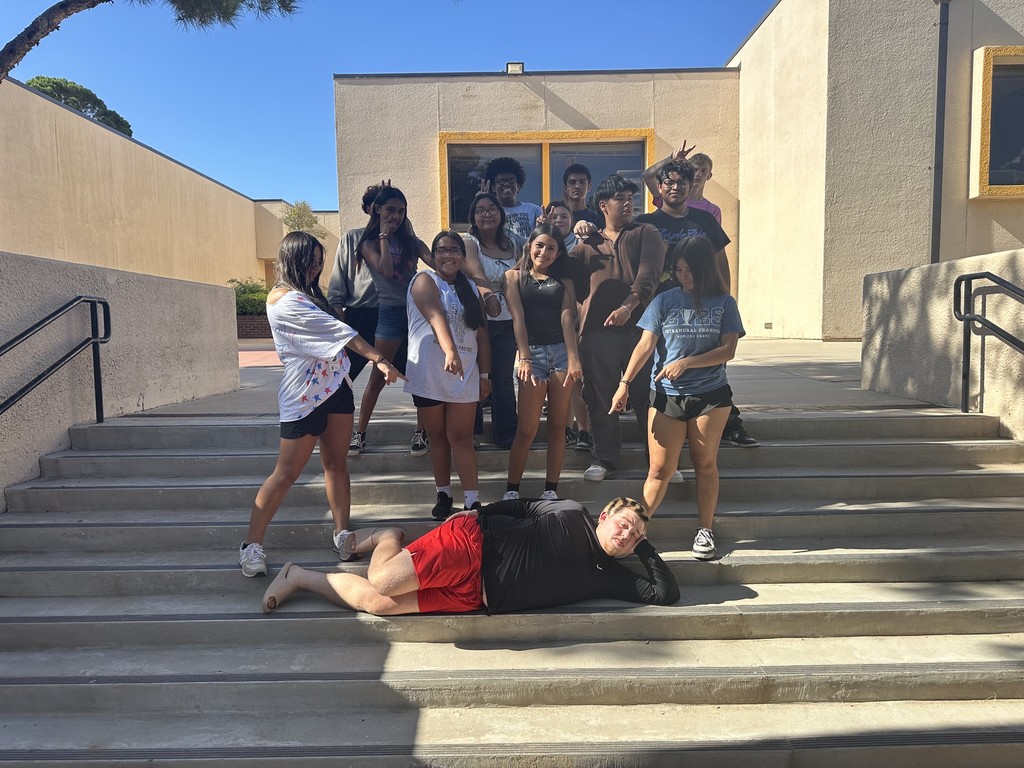 Students stand on the steps of the courtyard pointing at another student reclining in front of them.