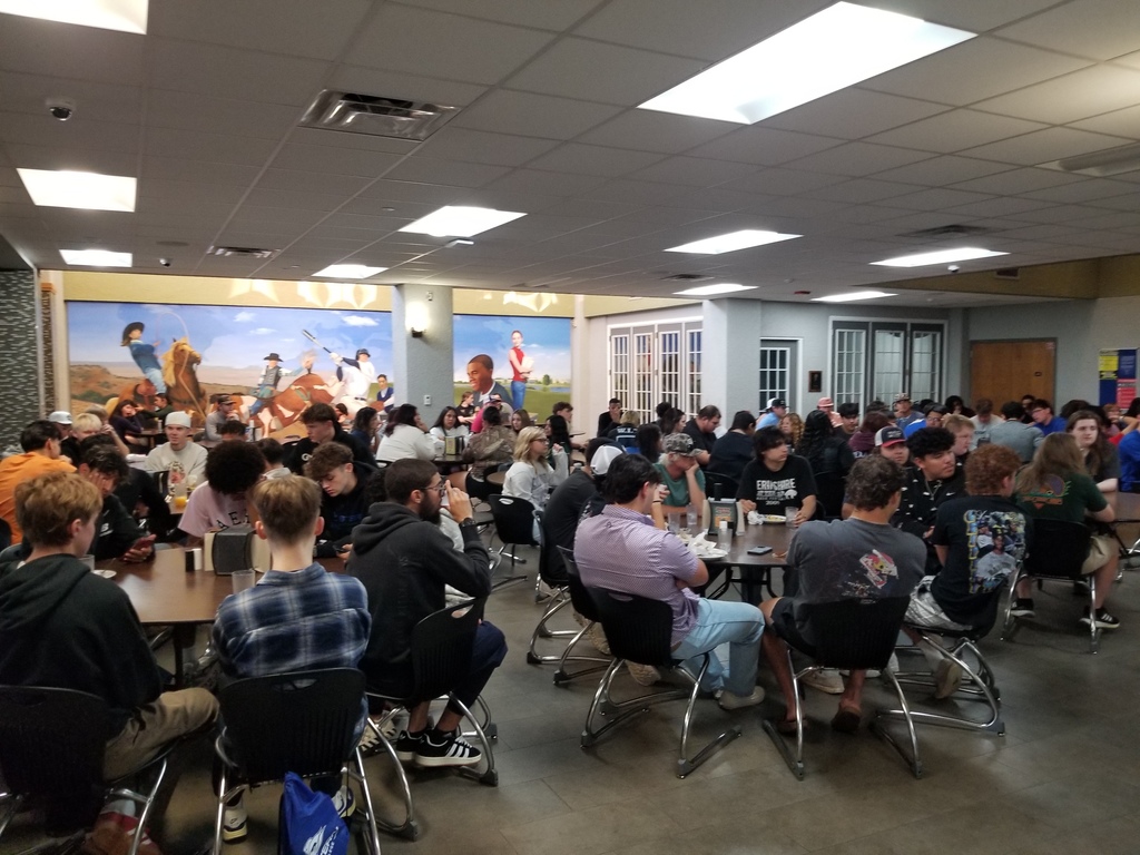 Students sit at round tables in the Cafeteria listening to Mr. Ramon speak.