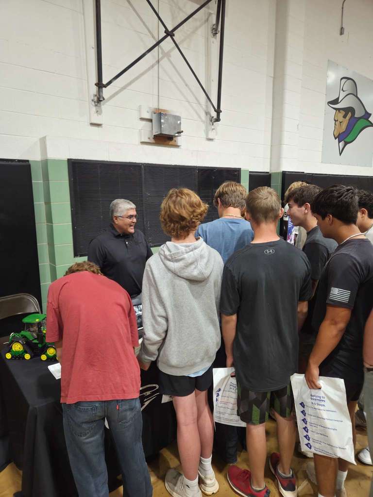 A group of students gathers around a booth featuring farm equipment at the college fair, speaking with a representative.