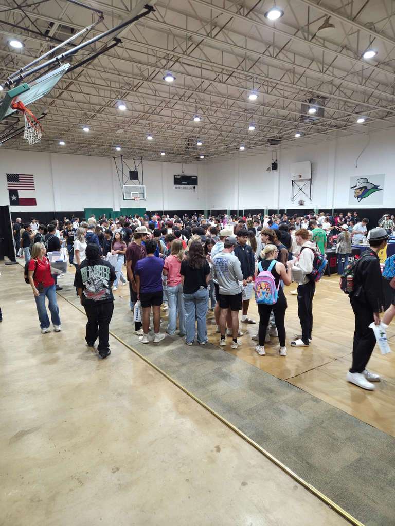 Wide view of a packed college fair in a gymnasium, with many students visiting booths and filling the space.