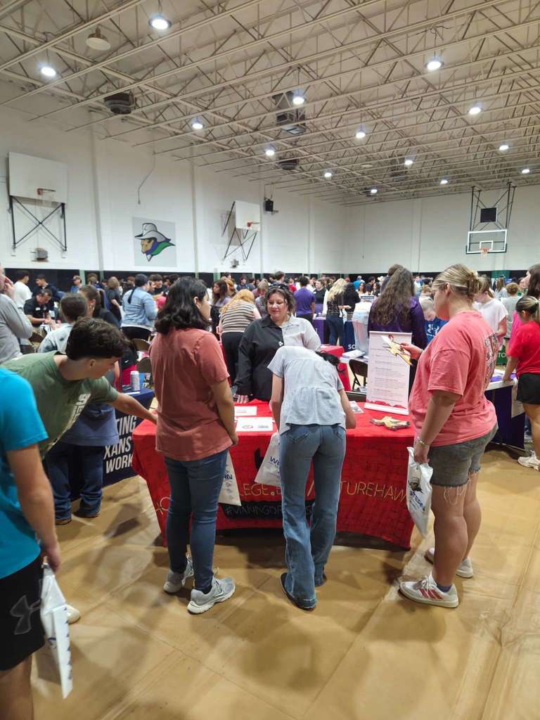 Students crowd around a red-covered college booth at the fair, asking questions and collecting brochures.
