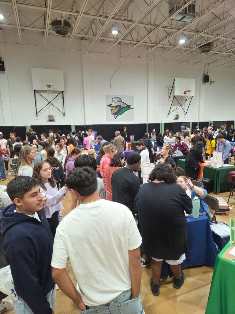 A busy college fair with students standing in groups, talking to representatives at booths set up in the gym.