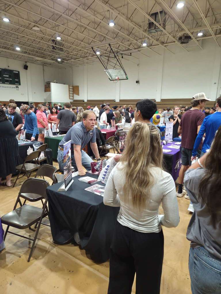 A college fair scene with a representative leaning on a booth table, speaking with a group of students in a crowded gym.