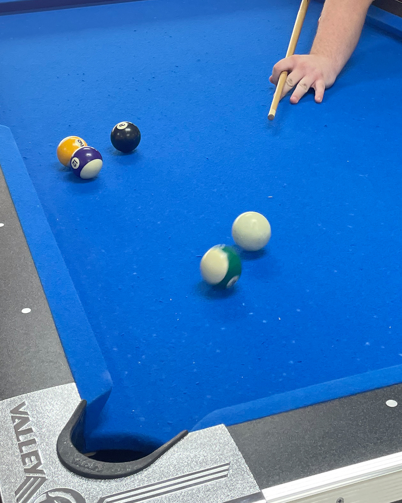 A person plays pool on a blue felt table, holding a cue stick while two balls collide near a corner pocket.