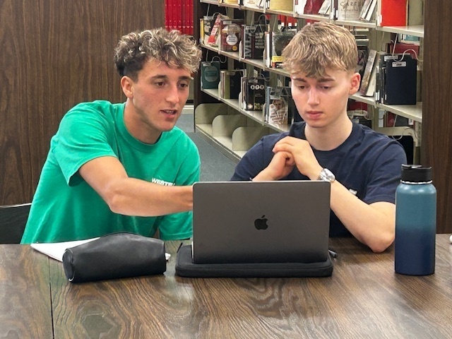 A tutor in a green shirt helps a student while they look at a laptop screen.