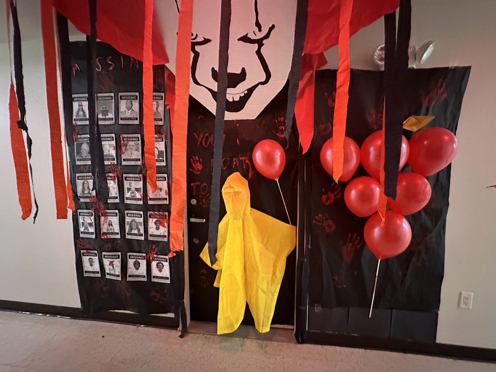 Pennywise-themed door with red balloons, “Missing” posters, red streamers, and yellow raincoat figure decoration.