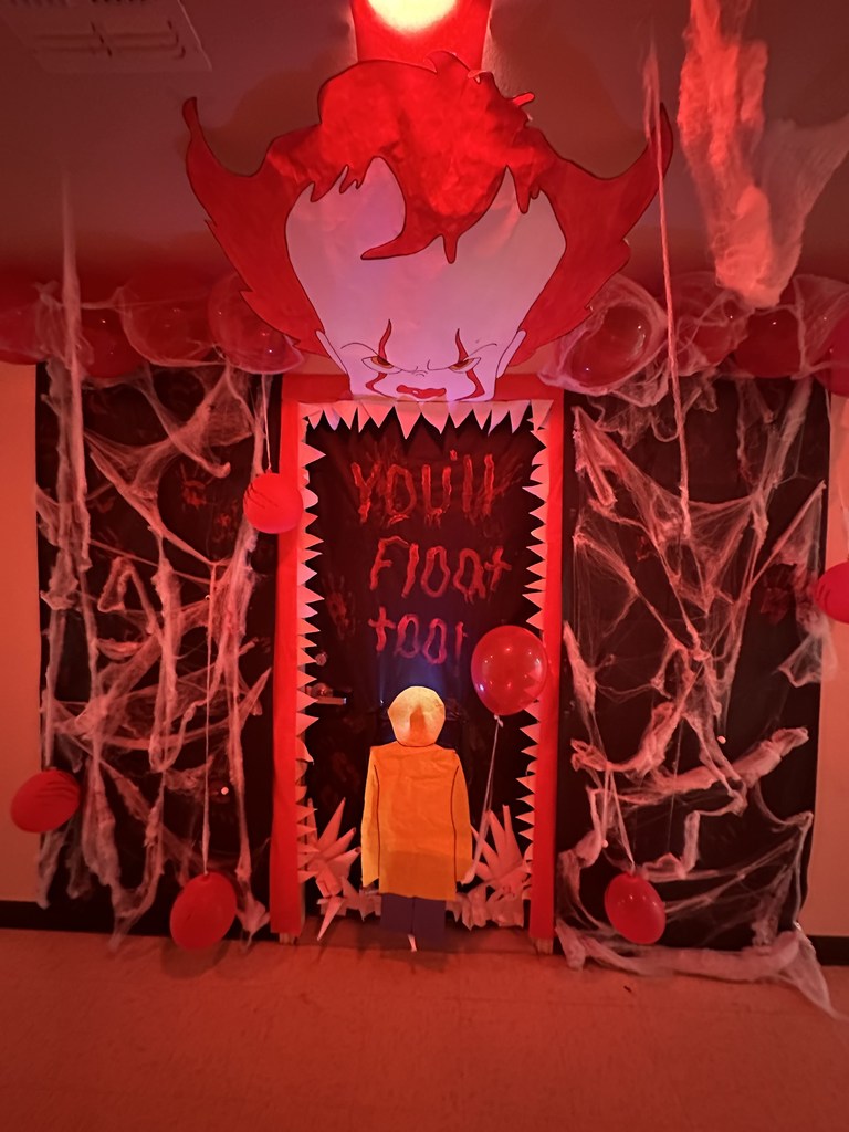 Hallway door decorated with Pennywise clown face, cobwebs, red balloons, and “You’ll float too” in red letters.