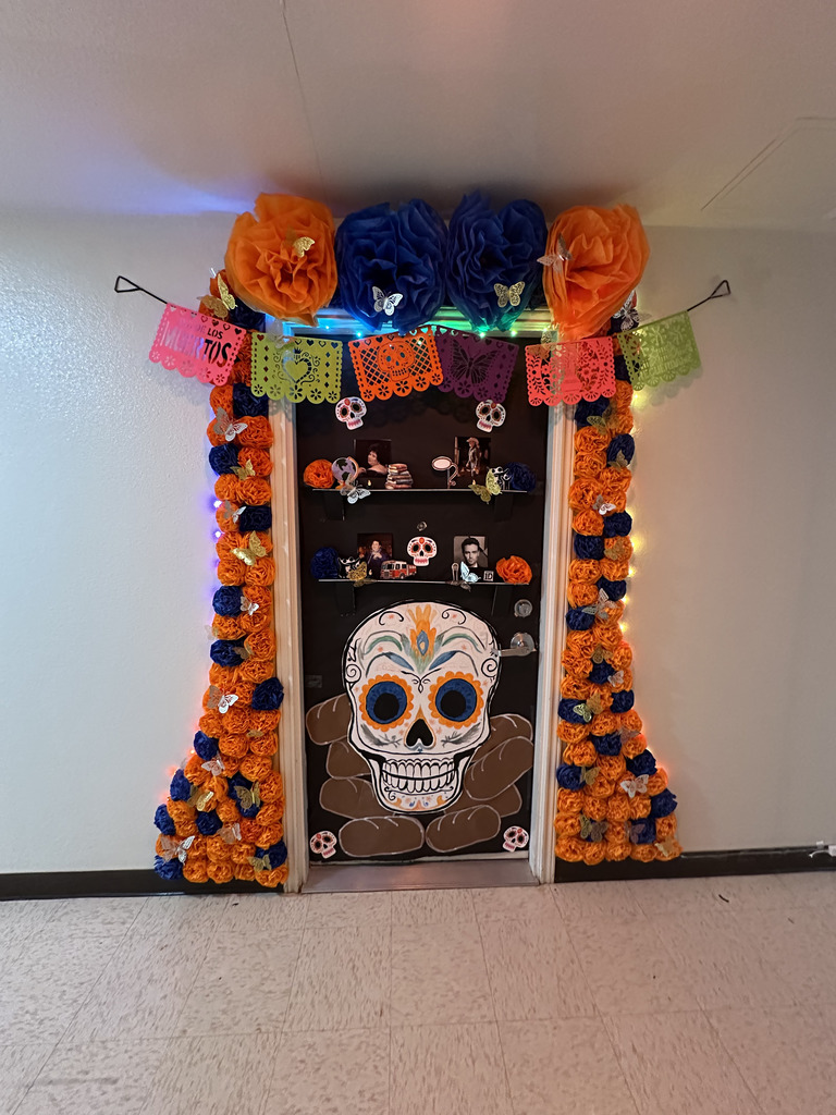 Door decorated for Día de los Muertos with colorful flowers, papel picado, sugar skull, and family photos.