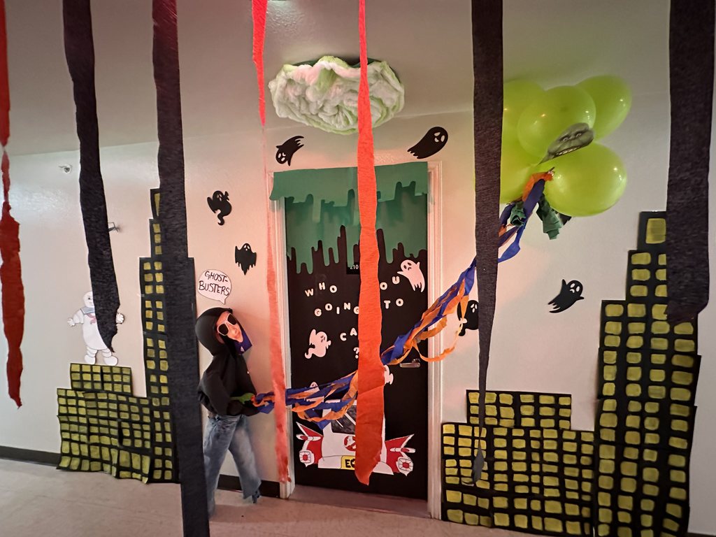 Ghostbusters-themed door with green balloons, cityscape cutouts, ghosts, and a child in costume holding streamers.
