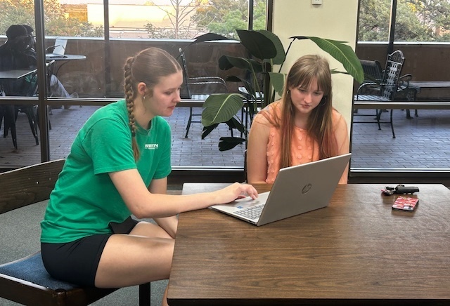 Two students at a table with a laptop, studying near large windows in the Western Texas College Library.