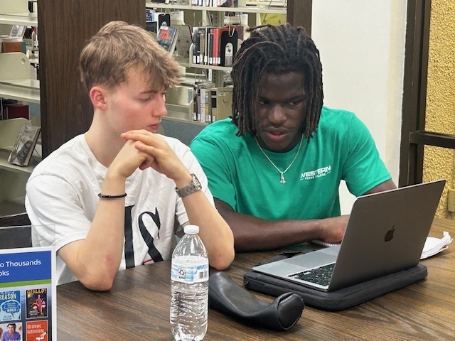 Two students studying together in the library, one wearing a green Western Texas College shirt, working on a laptop.