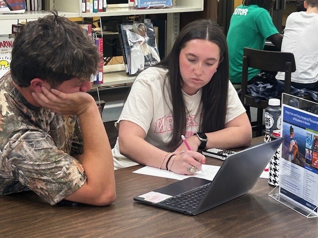 A tutor helps another student in the library, writing notes beside an open laptop at Western Texas College.