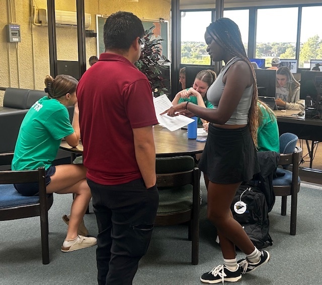 Students collaborating in the library, reviewing papers while others study at computers.