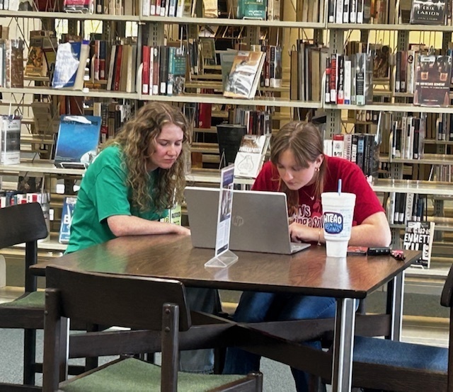 A tutor helps a student studying at a library table, focused on a laptop.