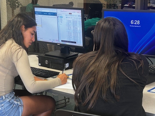 Two students working together at desktop computers, one taking notes while studying.