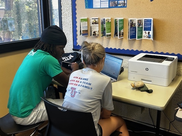 A tutor works with a student at a desk with a laptop and printer nearby.