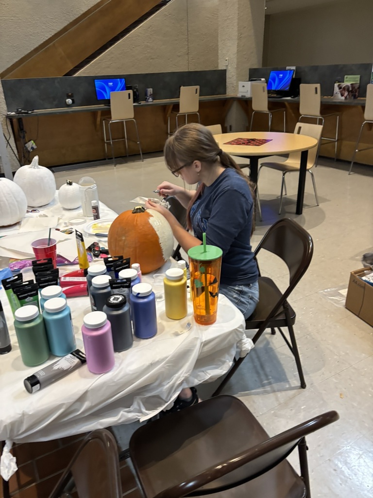 Student focused on painting a large orange pumpkin with art supplies nearby.