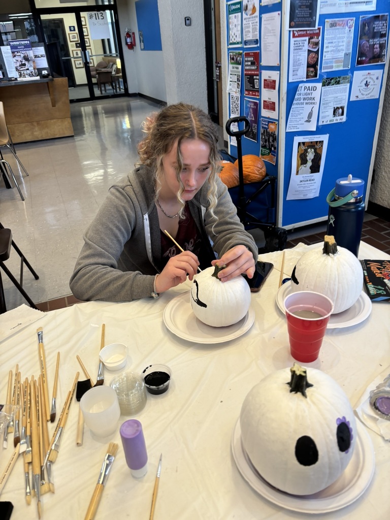 Student carefully paints a white pumpkin with brushes and supplies on the table.