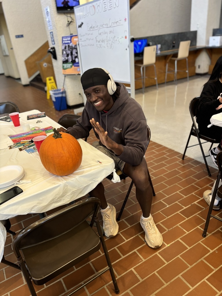 Student smiling and waving while sitting at a table with a pumpkin during the decorating contest.