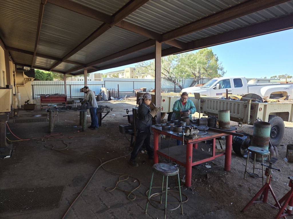 Students work on welding projects outdoors under a covered area with tools, tables, and equipment nearby.