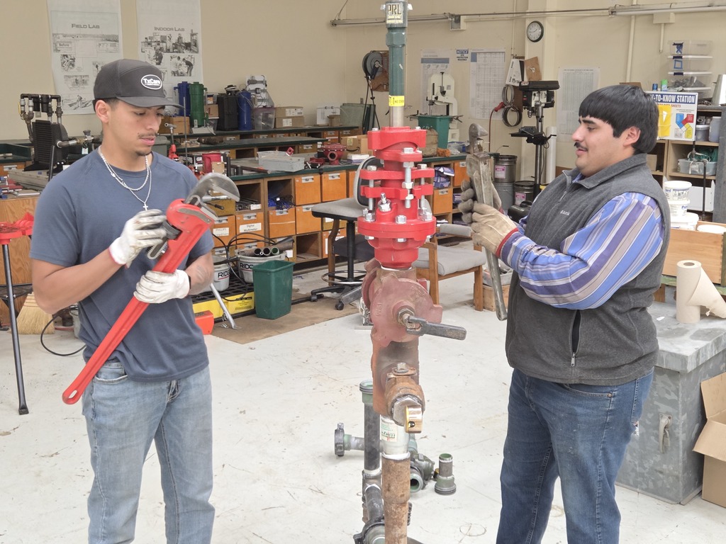 Two students use wrenches to tighten parts on an oilfield training assembly.