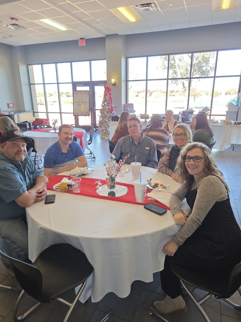 Faculty and staff seated at a decorated round table during the holiday luncheon with large windows behind.