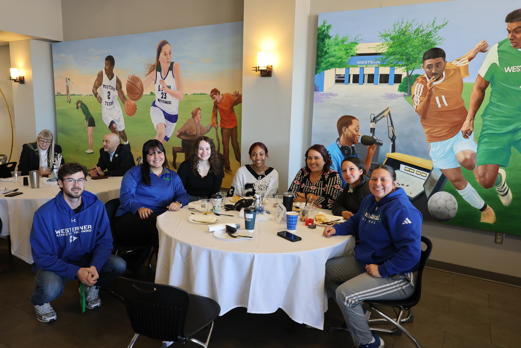 WTC Athletics' coaches smile while sitting at a table covered in a white tablecloth in the cafeteria with a mural in the background.