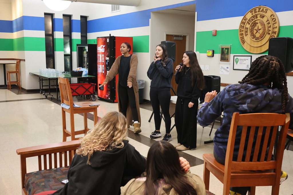 Students and coach sing karaoke together on stage in the WTC Student Commons with speakers and microphones.