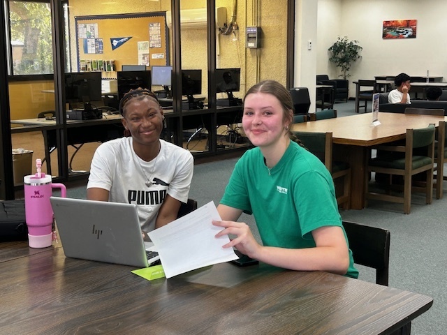 Two students smile while studying together at a table in the Western Texas College library, reviewing papers on a laptop.