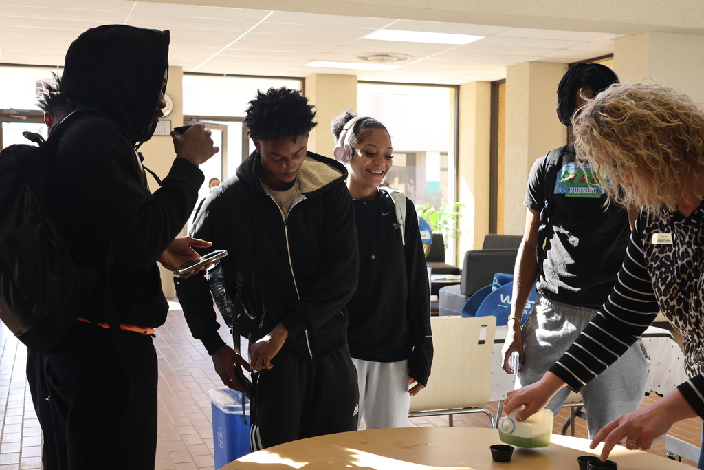 Three men and a woman smile while drinking from sample cups while another woman pours from a plastic jug.