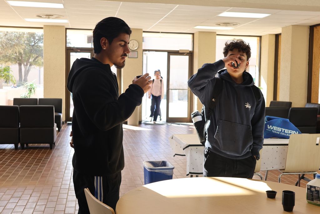 One man looks at a sample cup in his hand while another man drinks form a sample cup. standing next to a round table.