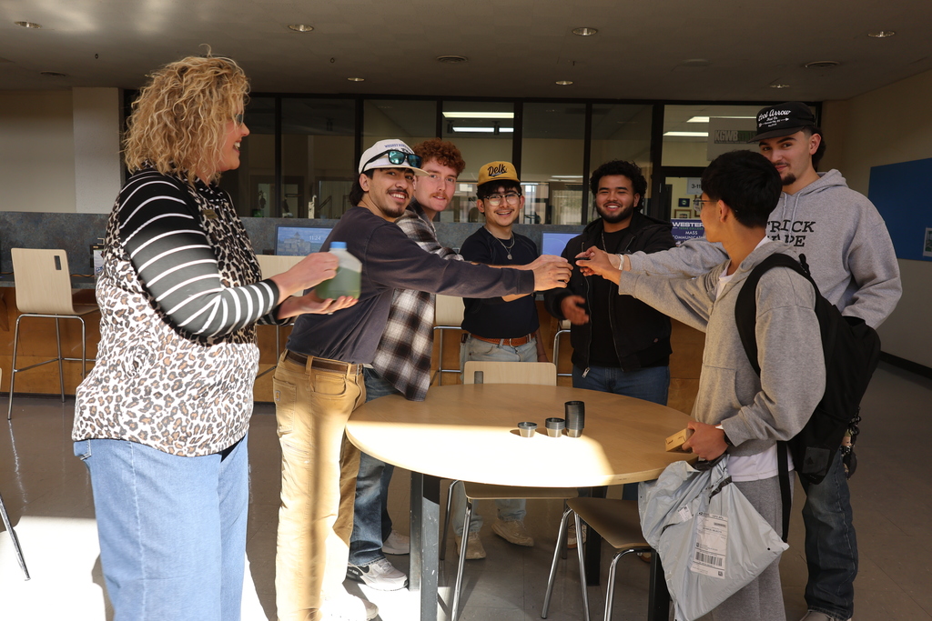 Men standing around a round table hold up sample cups together as a woman holding a jug watches.